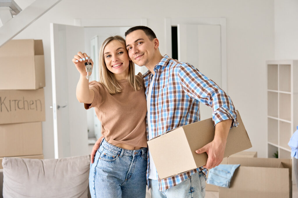 Happy young couple home owners holding keys in new home. Smiling independent millennial man and woman first time homeowners carrying boxes on moving day. Mortgage loan, new house ownership concept.