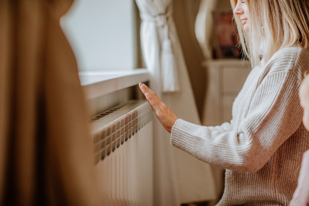 Young woman in long winter beige sweater is posing at home near the radiator